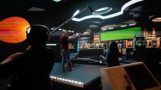 《OG官网》的导演杰森·墨菲站在一个虚拟宇宙飞船的舞台上. 他正在和一名工作人员交谈，另一名工作人员拿着吊杆麦克风.